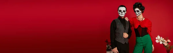 Élégant couple dia de los muertos en maquillage catrina regardant caméra près de fleurs sur rouge, bannière — Photo de stock