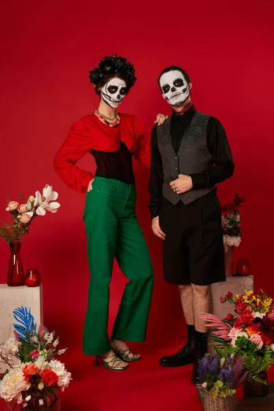 Elegante pareja en maquillaje de cráneo de azúcar cerca del tradicional día de los muertos ofrenda con flores sobre rojo - foto de stock