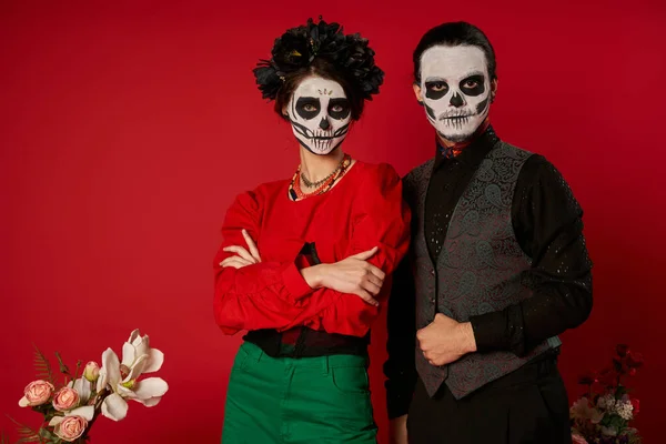 Casal em assustador dia de los muertos maquiagem e traje festivo olhando para a câmera perto de flores no vermelho — Fotografia de Stock
