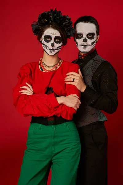Día de los muertos pareja en maquillaje espeluznante, hombre abrazando a mujer con los brazos cruzados en rojo, pancarta - foto de stock
