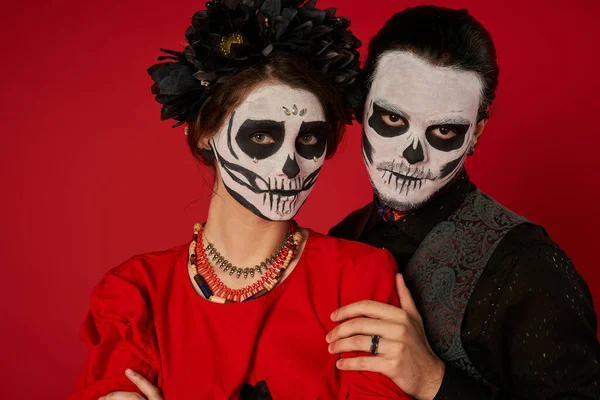 Stylish couple in scary sugar skull makeup looking at camera on red, dia de los muertos festival — Stock Photo