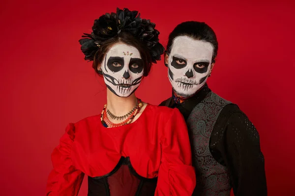 Retrato de pareja espeluznante en dia de los muertos maquillaje de cráneo de azúcar mirando a la cámara sobre fondo rojo - foto de stock