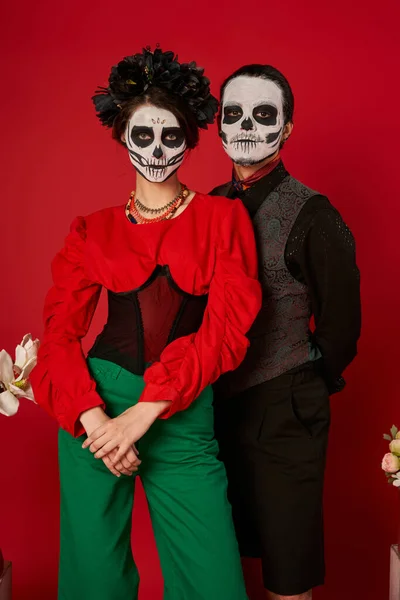 Couple in festive attire and catrina makeup posing on red backdrop, dia de los muertos celebration — Stock Photo