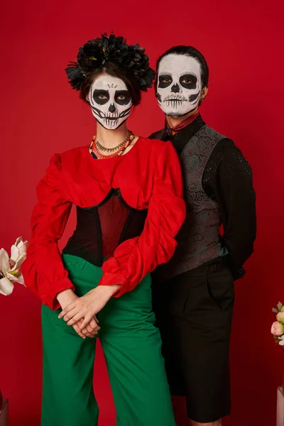 Pareja en traje festivo y maquillaje de catrina posando sobre fondo rojo, día de los muertos celebración - foto de stock