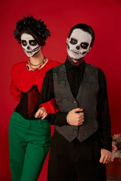 Young couple in scary skeleton makeup and stylish clothes posing on red, dia de los muertos festival — Stock Photo