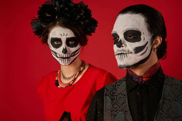 Dia de los muerts пара, жінка в макіяжі черепа і чорний вінок дивиться на камеру біля людини на червоному — стокове фото