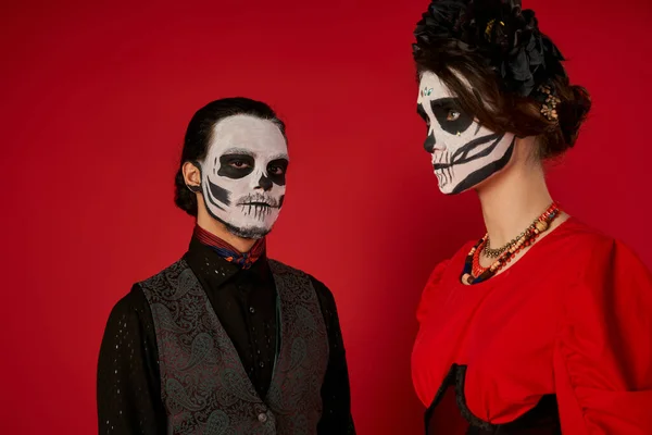 Uomo in trucco cranio guardando la fotocamera vicino alla donna in ghirlanda nera, dia de los muertos coppia su rosso — Foto stock