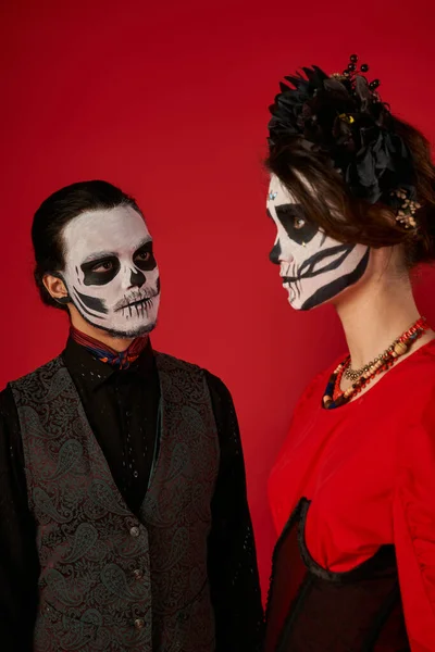 Homme effrayant dans le maquillage du crâne de sucre regardant la femme en couronne noire, dia de los muertos couple sur rouge — Photo de stock