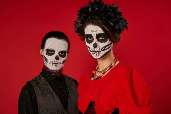 Eerie couple in traditional catrina makeup looking at camera on red, dia de los muertos celebration — Stock Photo
