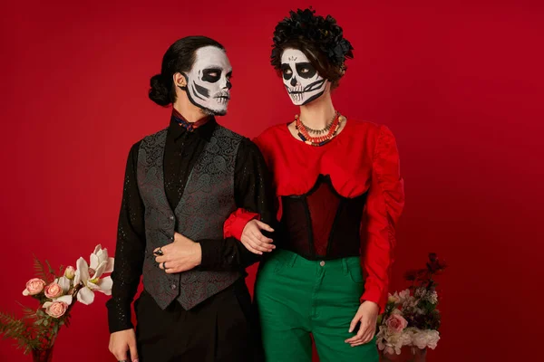 Stylish couple in skeleton makeup looking at each other near flowers on red, dia de los muertos fest — Stock Photo
