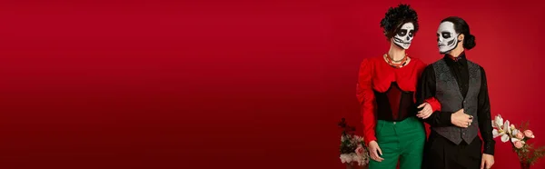 Elegant couple in catrina makeup posing near flowers on red, dia de los muertos festival, banner — Stock Photo