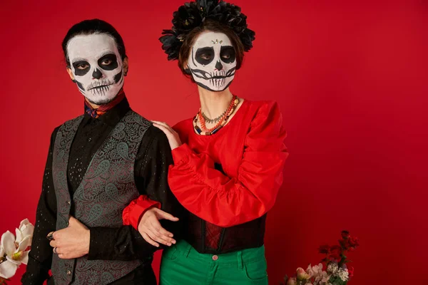 Stylish couple in traditional dia de los muertos makeup looking away near flowers on red backdrop — Stock Photo
