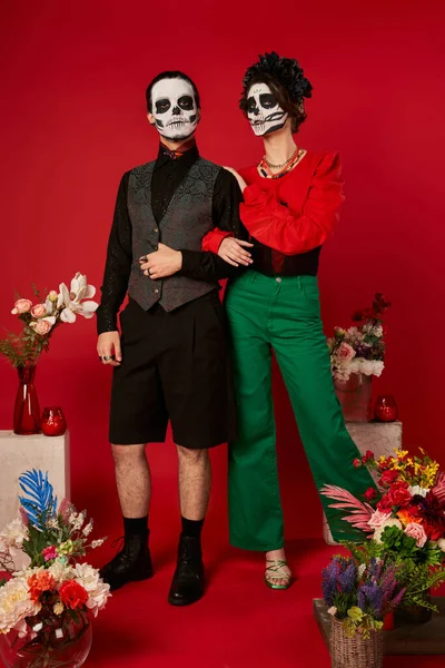 Toute la longueur du couple dans le maquillage du crâne près traditionnelle Jour de l'ofrenda mort avec des fleurs sur le rouge — Photo de stock