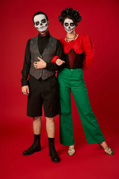 Piena lunghezza di coppia in abiti alla moda e trucco scheletro sul rosso, dia de los muertos celebrazione — Foto stock