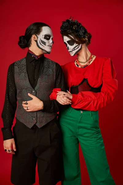 Couple in trendy attire and skeleton makeup on red, dia de los muertos celebration — Stock Photo