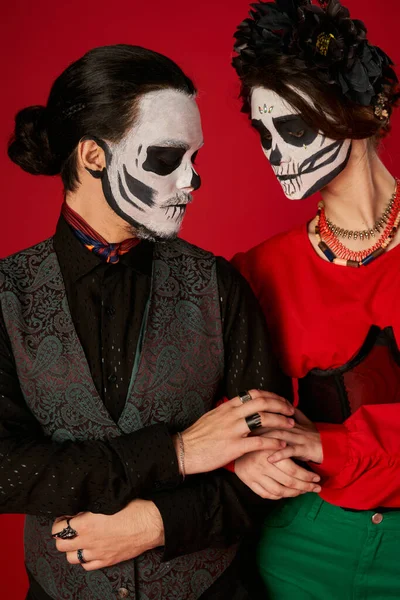 Trendy couple in sugar skull makeup touching hands of each other on red, dia de los muertos festival — Stock Photo