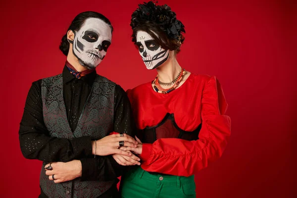 Casal em maquiagem crânio tocando as mãos uns dos outros e olhando para a câmera no vermelho, Day of Dead fest — Fotografia de Stock