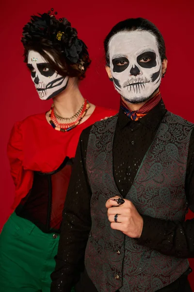 Homme dans le crâne maquillage regarder caméra près de la femme en couronne noire, dia de los muertos couple sur rouge — Photo de stock