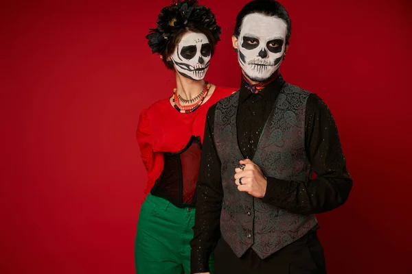 Man in catrina makeup looking at camera near woman in black wreath, dia de los muertos couple on red — Stock Photo