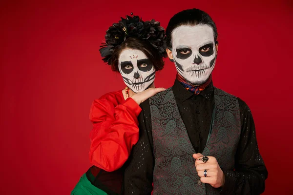 Woman in sugar skull makeup and black wreath leaning on shoulder of spooky man on red, Day of Dead — Stock Photo