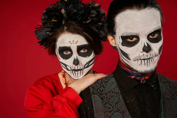 Hispanic culture, couple in dia de los muertos traditional makeup looking at camera on red, portrait — Stock Photo