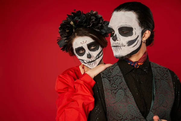 Woman in catrina makeup and black wreath leaning on shoulder of eerie man on red, Day of Dead — Stock Photo