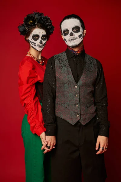 Stylish couple in dia de los muertos sugar skull makeup holding hands and looking at camera on red — Stock Photo