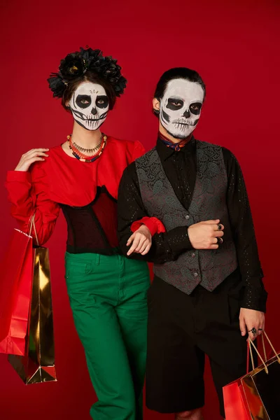Elegante pareja en día de los muertos maquillaje de pie con bolsas de compras en rojo, venta de temporada - foto de stock