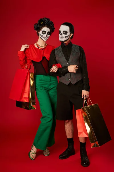 Longueur totale du couple dans dia de los muertos maquillage debout avec des sacs à provisions sur rouge, vente festive — Photo de stock