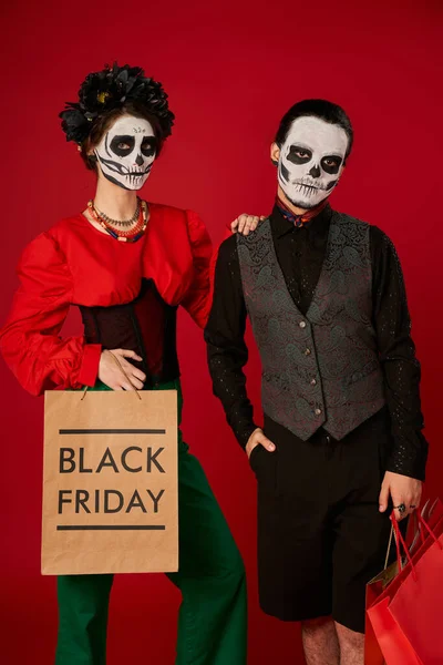 Paar in dia de los muertos Make-up, Frau mit schwarzer Freitagseinkaufstasche in der Nähe von gruseligen Mann auf rot — Stockfoto