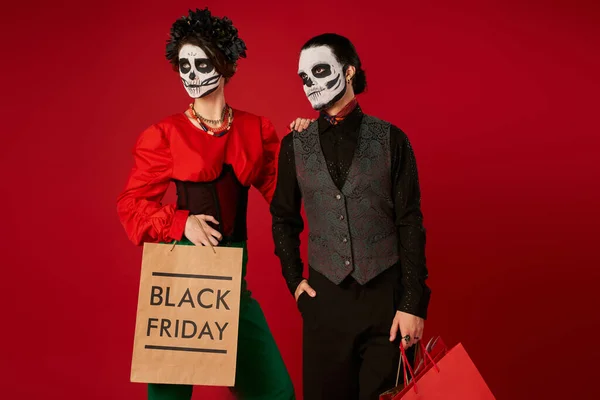 Coppia elegante in dia de los muertos cranio trucco in piedi con borse della spesa su rosso, venerdì nero — Foto stock