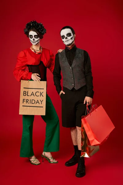 Полная длина модной пары в сахарном черепе макияж держа сумки для покупок на красный, черный пятница — стоковое фото