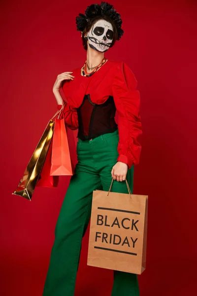 Donna alla moda in dia de los muertos trucco in posa con borse della spesa su rosso, venerdì nero vendita — Foto stock