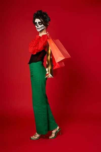 Longitud completa de la mujer en el Día de Muertos maquillaje y traje festivo posando con bolsas de compras en rojo - foto de stock
