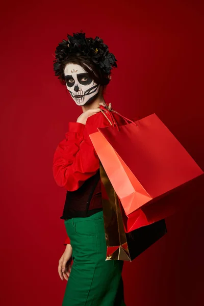 Femme élégante en couronne noire et maquillage catrina tenant des sacs à provisions et regardant la caméra sur rouge — Photo de stock