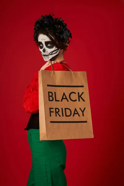 Giovane donna in dia de los muertos trucco e ghirlanda nera tenendo nero venerdì shopping bag su rosso — Foto stock