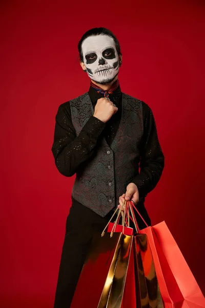 Man in scary sugar skull makeup and festive attire with shopping bags on red, seasonal sale — Stock Photo