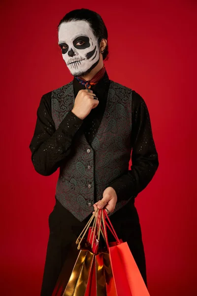 Hombre en día de los muertos maquillaje y elegante traje posando con bolsas de compras en rojo, venta de temporada - foto de stock