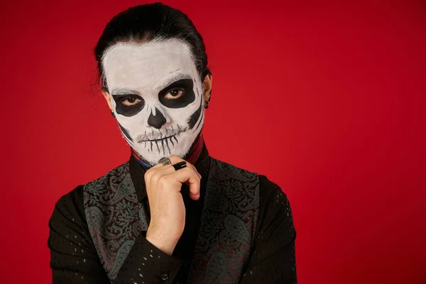 Hombre en espeluznante esqueleto maquillaje de la mano cerca de la barbilla y mirando a la cámara en rojo, Día de Muertos - foto de stock