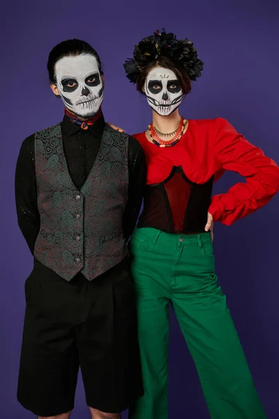 Стильная пара в праздничной одежде и dia de los muertos макияж глядя на камеру на синем фоне — стоковое фото
