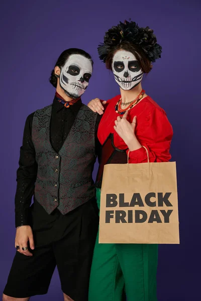 Donna in spaventoso cranio trucco e ghirlanda nera in possesso di Venerdì nero shopping bag vicino all'uomo su blu — Foto stock