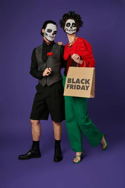 Woman in eerie catrina makeup posing with black friday shopping bag near spooky man on blue — Stock Photo