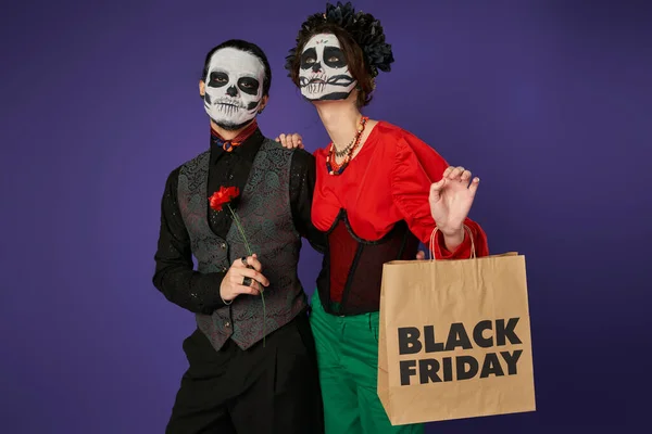 Donna spaventosa con nero venerdì shopping bag guardando lontano vicino all'uomo in zucchero cranio trucco su blu — Foto stock