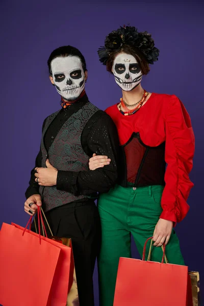 Stylish dia de los muertos couple in skull makeup posing with shopping bags on blue, seasonal sale — Stock Photo