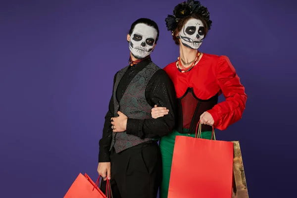 Елегантна пара в діа-де-лос-муарі черепа макіяж позує з сумками на синьому, сезонному розпродажі — стокове фото