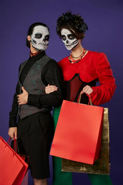 Coppia in dia de los muertos trucco e abbigliamento festivo che tiene borse della spesa su blu, vendita stagionale — Foto stock