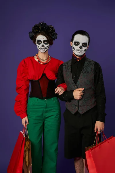 Dia de los muertos couple in sugar skull makeup holding shopping bags and looking at camera on blue — Stock Photo