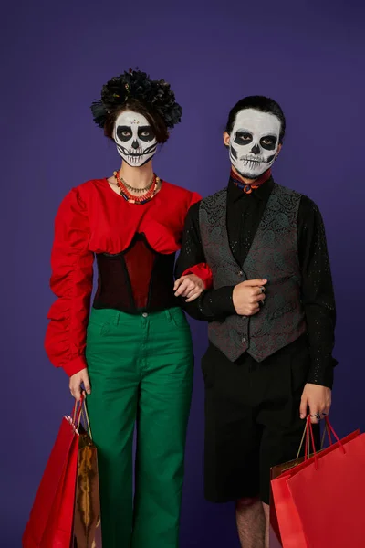 Dia de los muertos couple in sugar skull makeup holding shopping bags and looking at camera on blue — Stock Photo