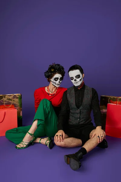 Dia de los muertos couple dans le maquillage squelette effrayant assis près de sacs à provisions sur bleu, vente saisonnière — Photo de stock