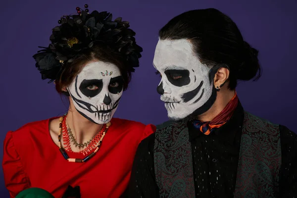 Woman in sugar skull makeup and black wreath looking at camera near spooky man on blue, Day of Dead — Stock Photo